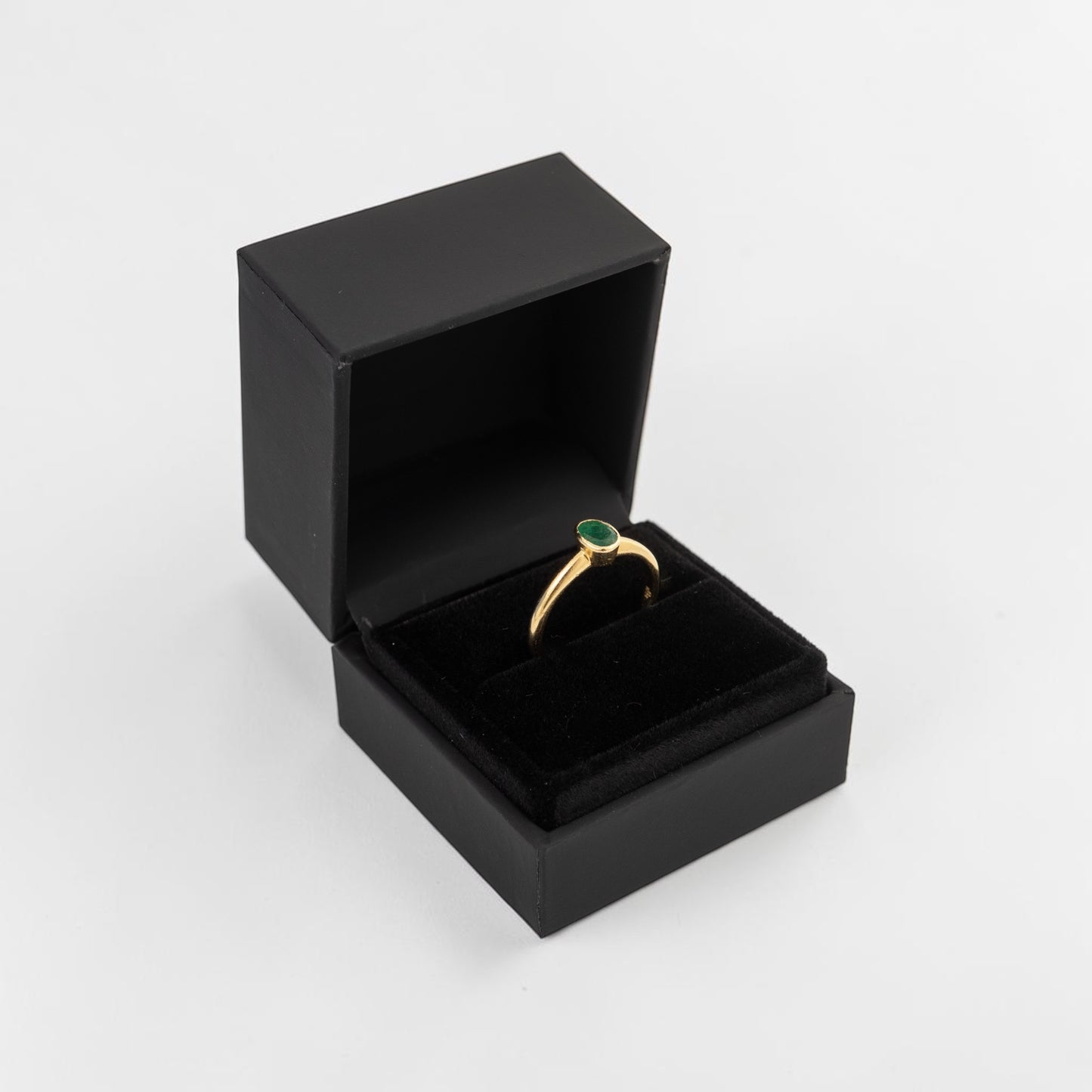 A gold ring with a small green oval gemstone, presented in an open black velvet jewelry box on a light gray background.