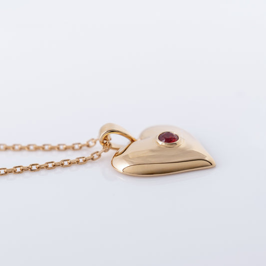 A polished gold heart pendant necklace with a small round red gemstone set in the center, resting on a clean white surface.