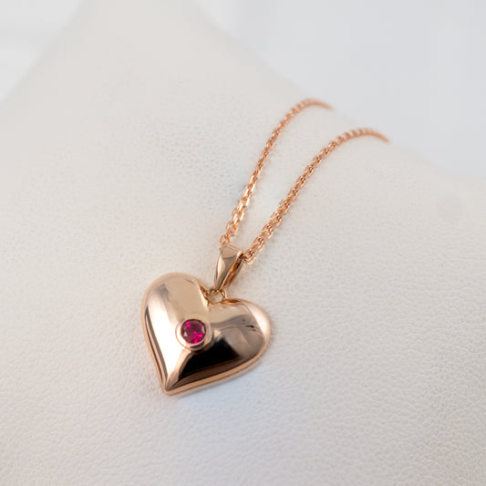 Rose gold heart-shaped pendant with a red gemstone on a light gray background