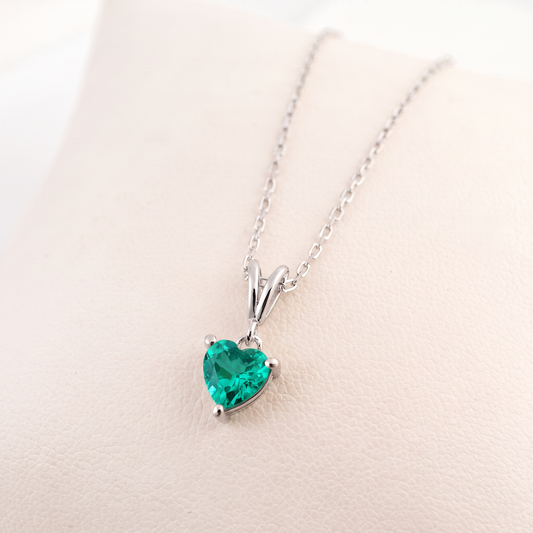 A heart-shaped emerald necklace pendant in a silver setting on a delicate silver chain, displayed on a light beige textured surface.