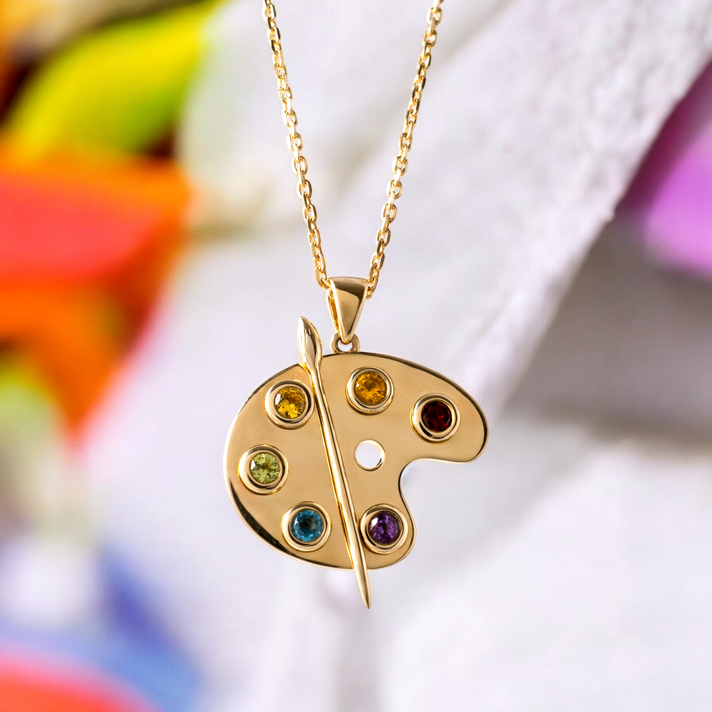 Gold artist palette pendant featuring vibrant gemstones displayed against a creative artistic background with paintbrushes, colours, and an easel, highlighting its unique design