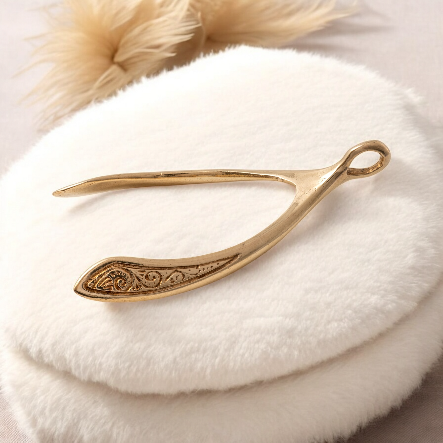 Solid gold wishbone pendant with intricate scroll detail displayed on a cream background, with a soft brown feather in the background.