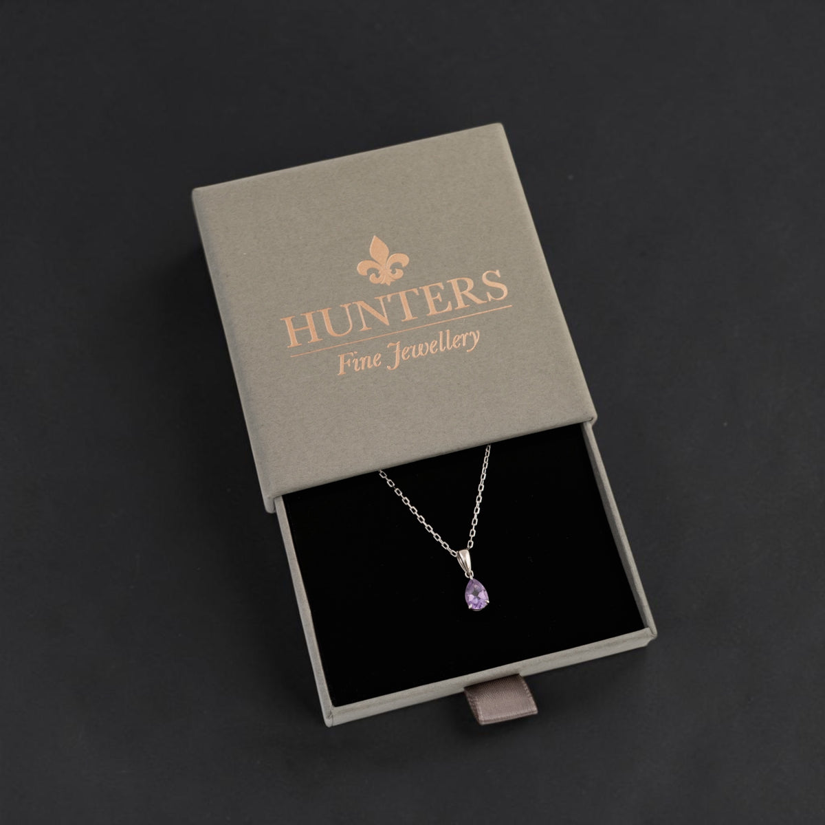 A silver pendant necklace with a teardrop-shaped purple gemstone displayed inside an open gray presentation box. The box lid features the brand name 'HUNTERS Fine Jewellery' in rose gold lettering.