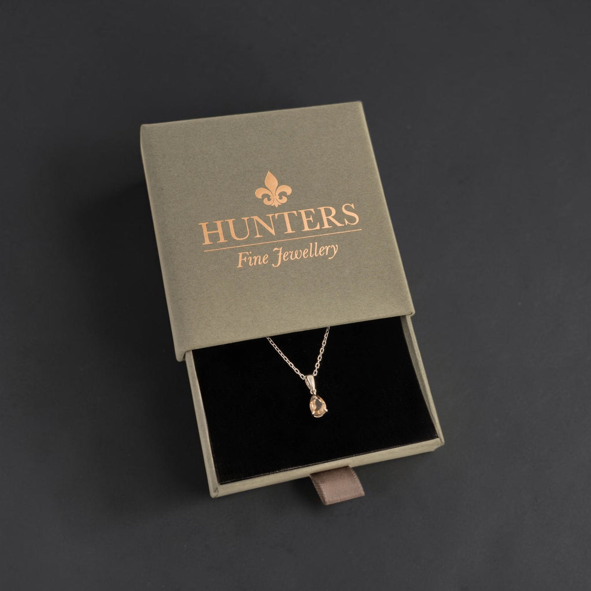 A silver-toned pendant necklace with a small citrine teardrop gemstone displayed inside an open Hunters Fine Jewellery box on a dark grey background.
