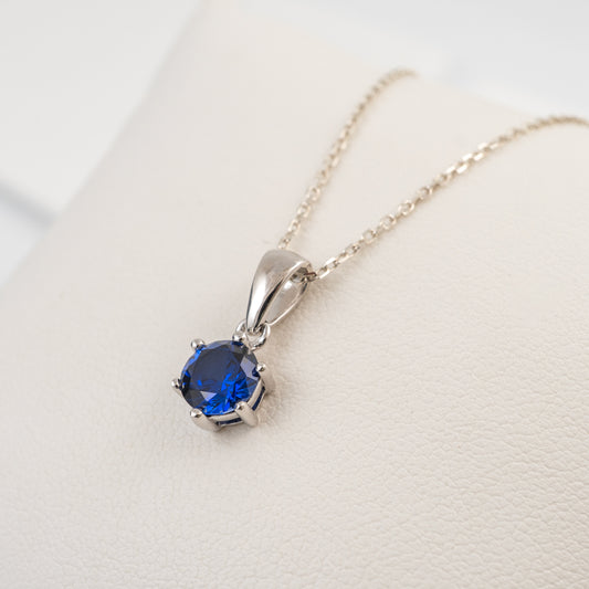 A silver lab grown sapphire necklace with a round blue gemstone in a prong setting, resting on a textured cream-colored surface.