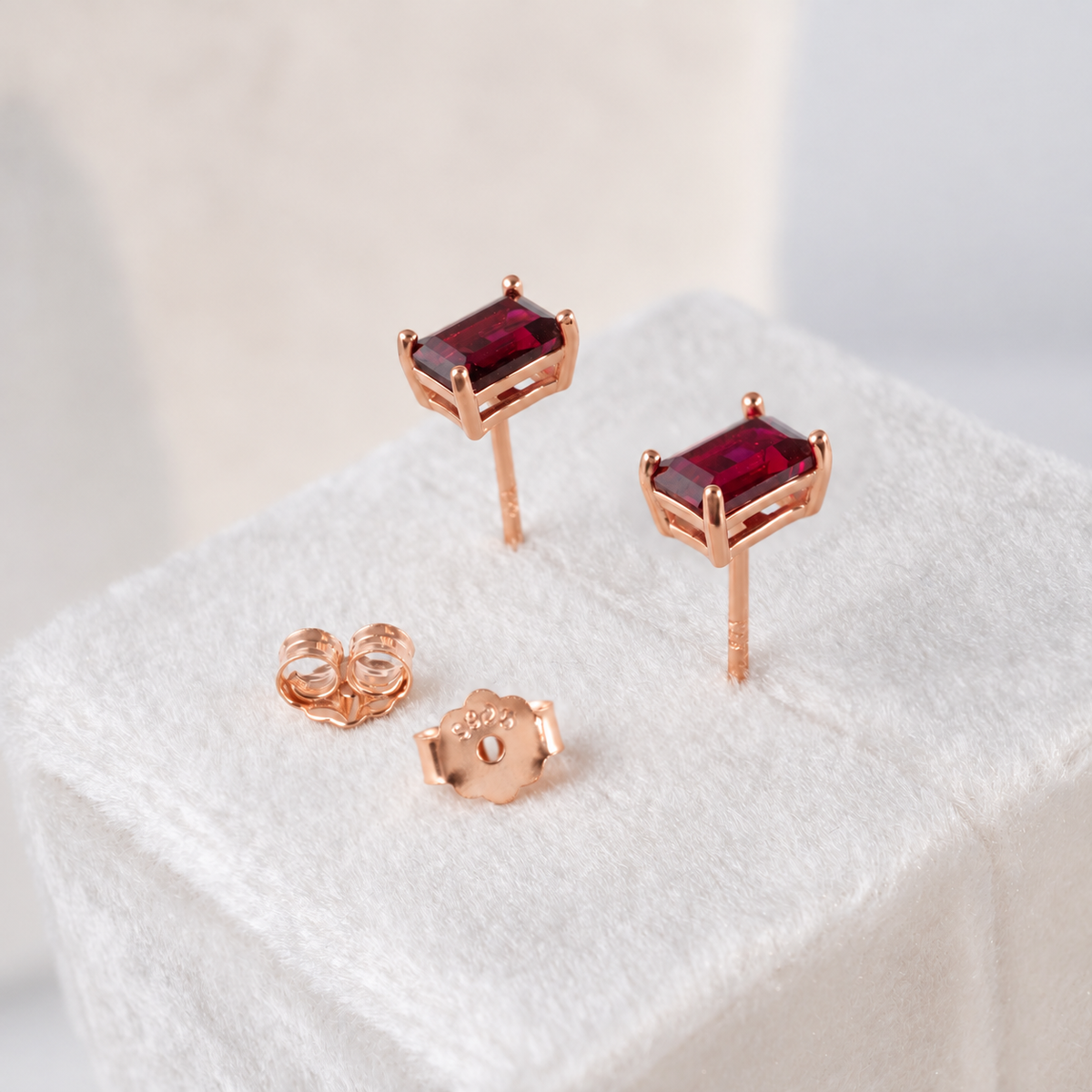 A pair of rose gold stud earrings with rectangular deep red gemstones, shown with their rose gold butterfly backings on a white textured display block.