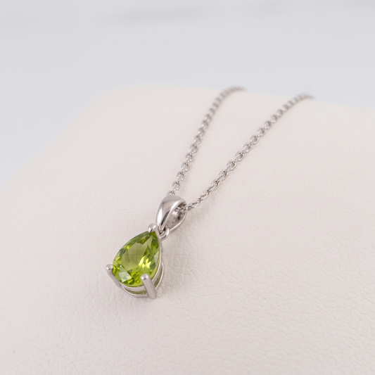 Angled view of a natural peridot teardrop necklace in sterling silver, showing the green pear-cut gemstone, claw setting and tapered bail on a white cushion.
