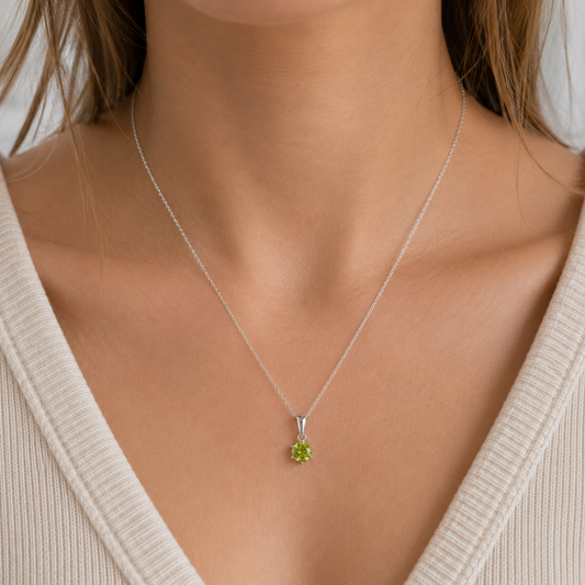 A close-up of a person's neck and chest wearing a delicate silver chain necklace with a small green gemstone pendant, paired with a cream-colored ribbed V-neck top.