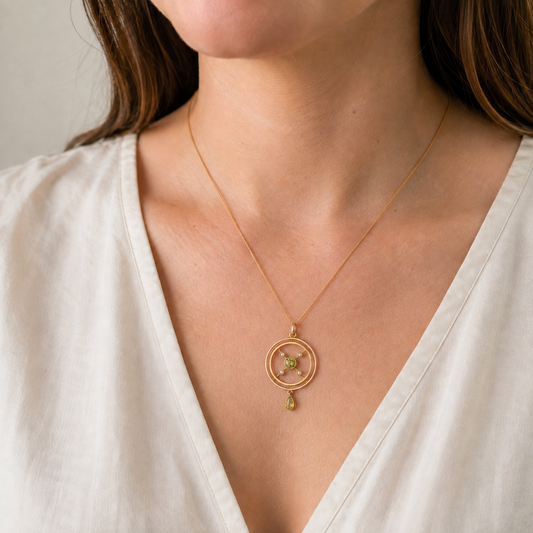 A close-up of a model wearing a delicate gold necklace with a circular pendant and a small green teardrop charm, paired with a white V-neck top.