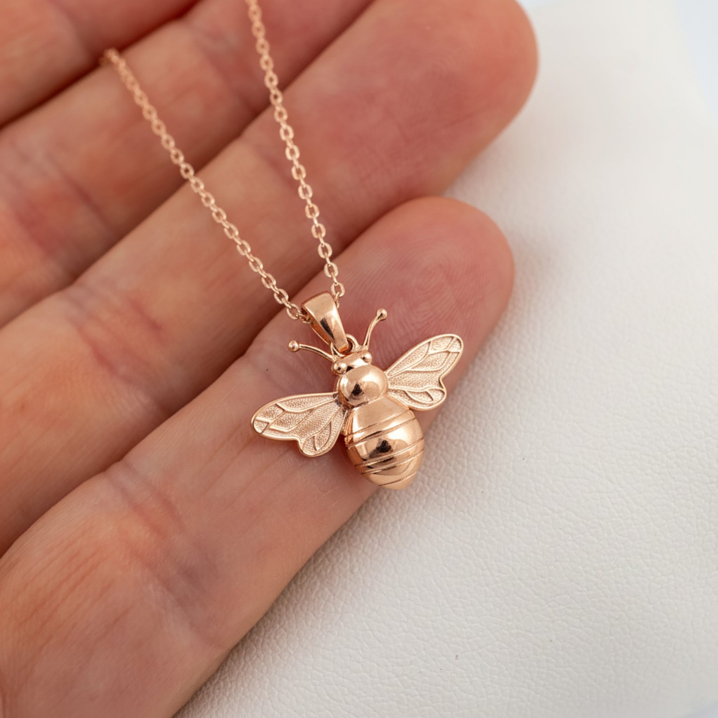 Rose gold bee pendant on a chain held between fingers against a white background