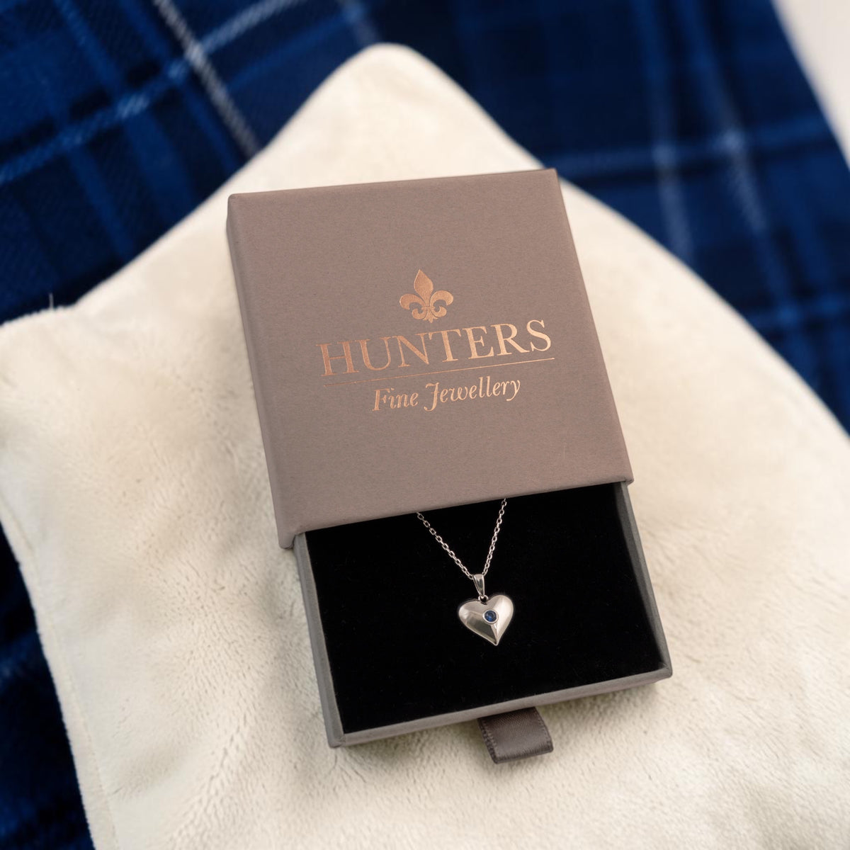 A silver heart pendant necklace displayed in an open gray Hunters Fine Jewellery gift box, resting on a white textured fabric with a blue plaid background.