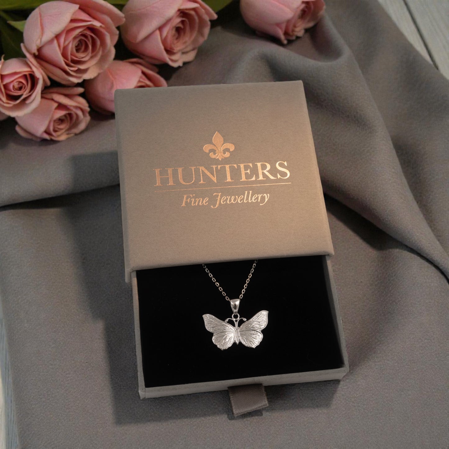 Butterfly necklace in a Hunters Fine Jewellery box with pink roses in the background