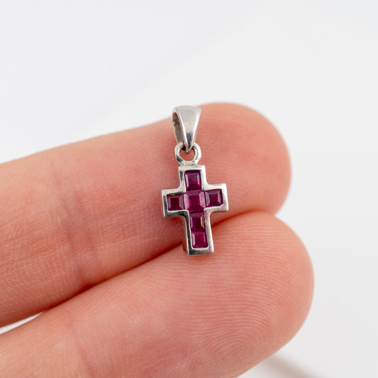 A white gold ross pendant with a central grid of small pinkish-red gemstones, held between two fingers to show its scale against a plain white background.