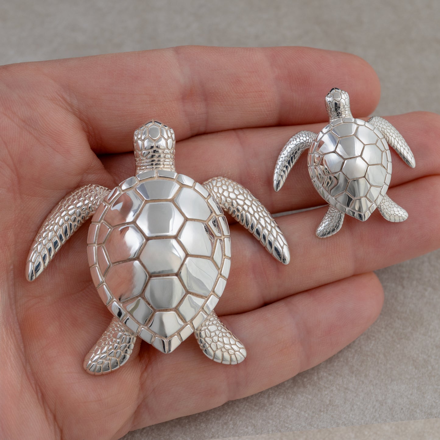 a samll and a large silver turtle figurines held in a hand against a neutral background