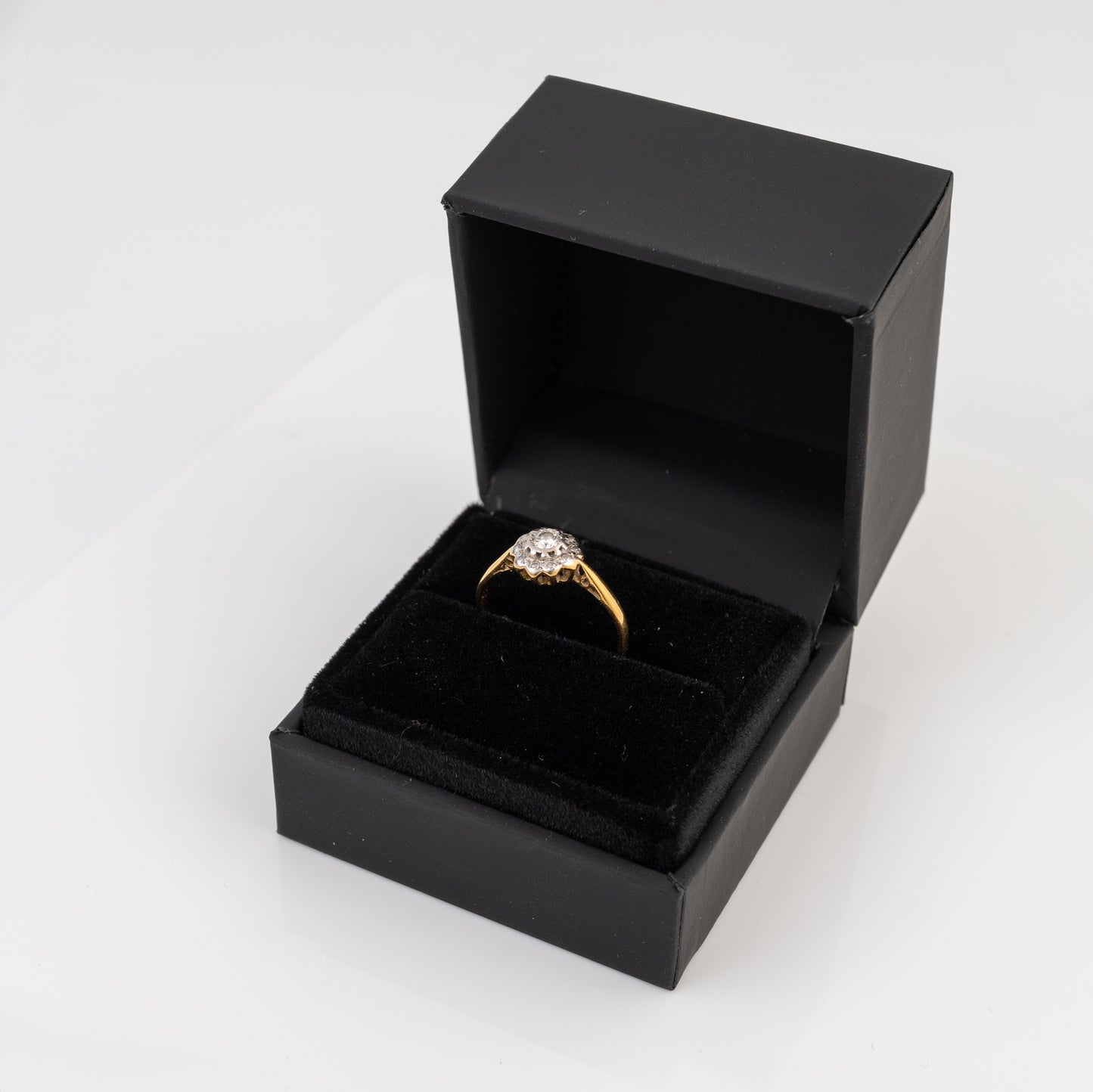A yellow gold diamond ring with a round clear gemstone displayed inside an open black presentation box with a black velvet insert, set against a plain white background.