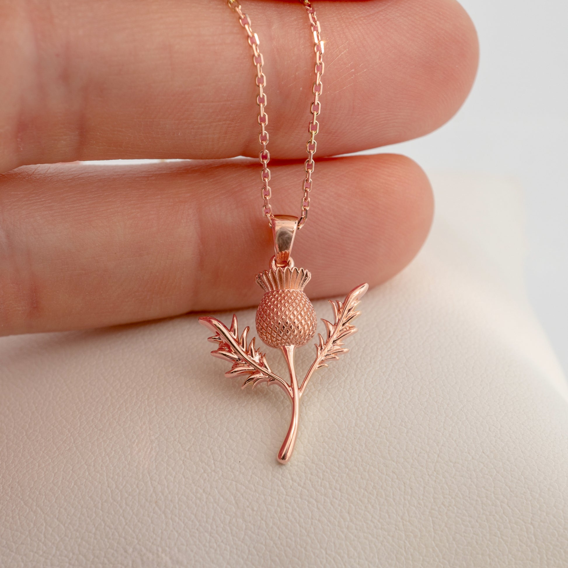 Rose gold thistle pendant necklace held between fingers on a light background