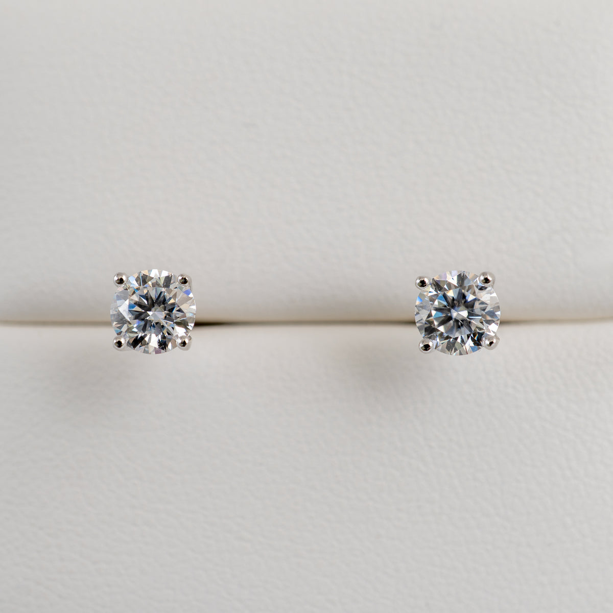 A pair of round-cut moissanite stud earrings in a silver-toned setting, displayed on a light textured surface.