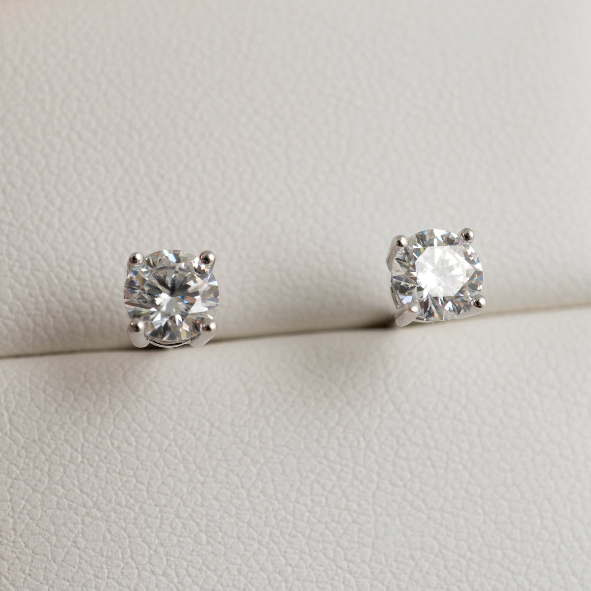 A pair of round-cut moissanite solitaire stud earrings in a silver-toned setting, displayed on a textured off-white surface.