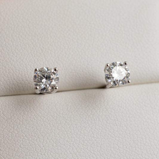 A pair of round-cut moissanite solitaire stud earrings in a silver-toned setting, displayed on a textured off-white surface.