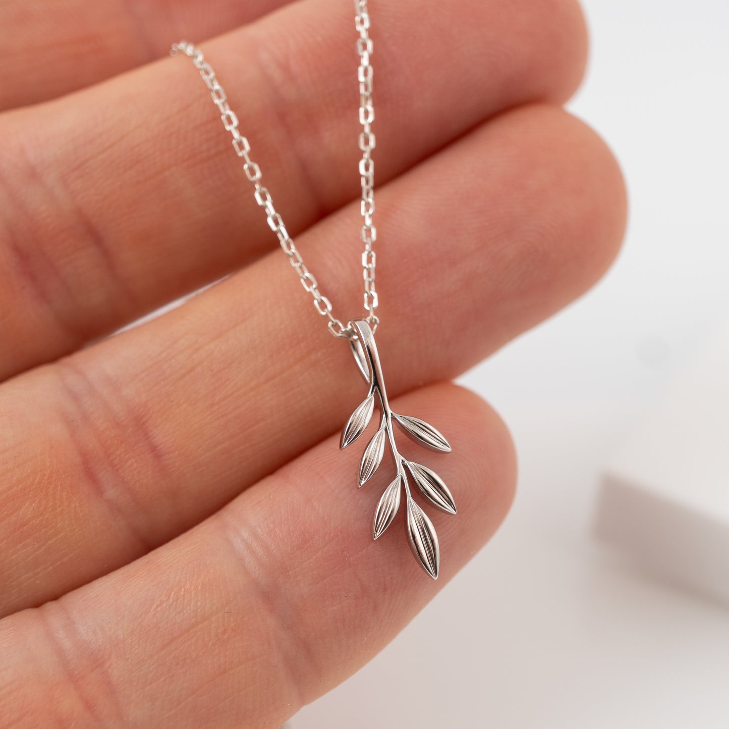 Silver olive leaf necklace held between fingers against a neutral background