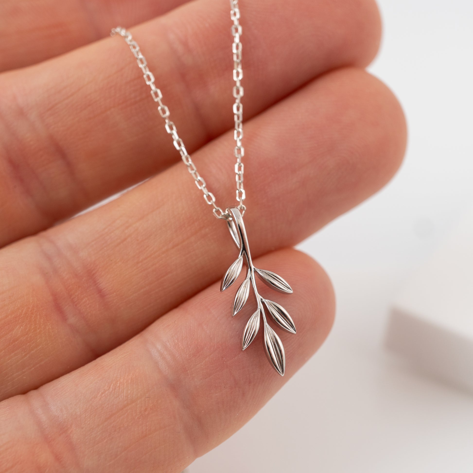 Silver olive leaf necklace held between fingers against a neutral background