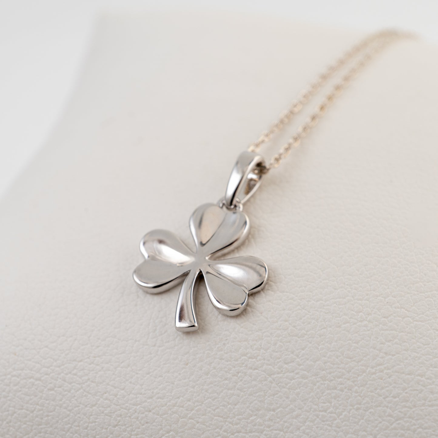 A polished silver three leaf shamrock pendant necklace on a delicate gold-tone chain, displayed on a textured cream-colored surface.