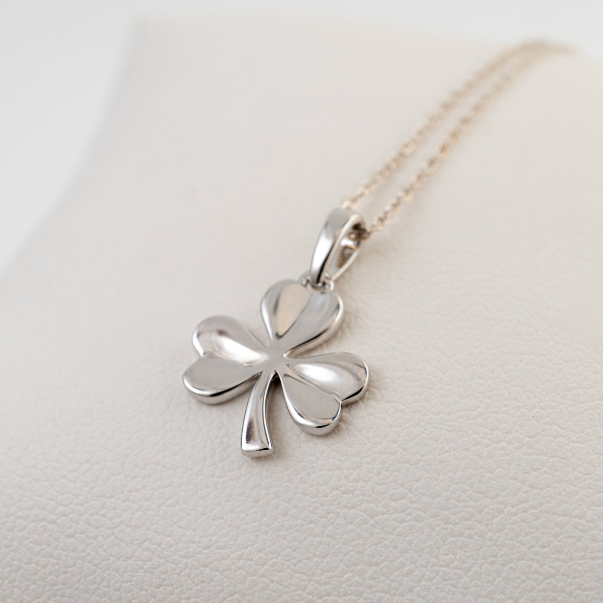 A polished silver three leaf shamrock pendant necklace on a delicate gold-tone chain, displayed on a textured cream-colored surface.
