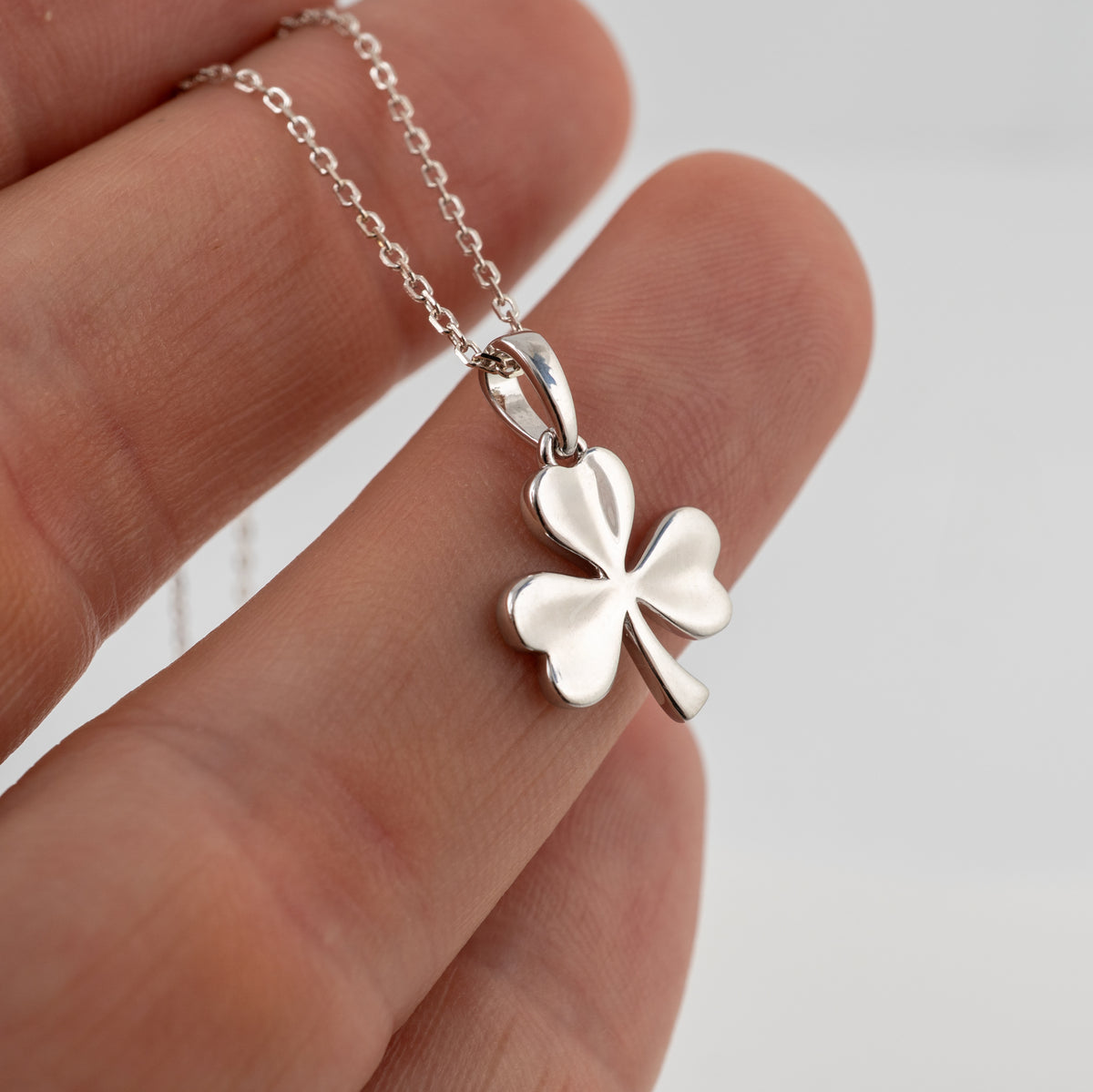 A silver shamrock necklace pendant necklace held on a person's fingers against a light gray background.