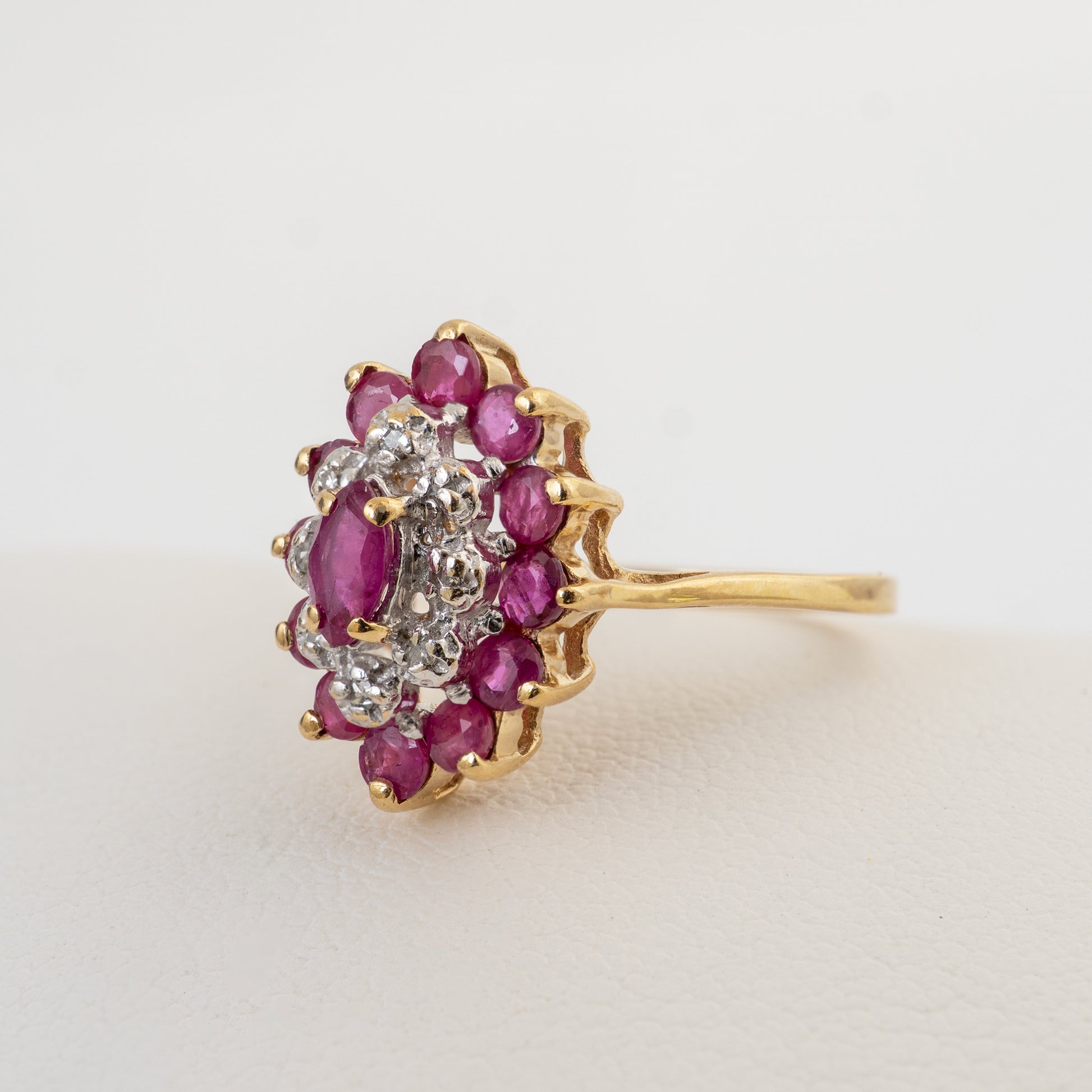 A yellow gold ring featuring a floral cluster setting with oval pinkish-red rubies and round clear diamonds, photographed on a light neutral background.