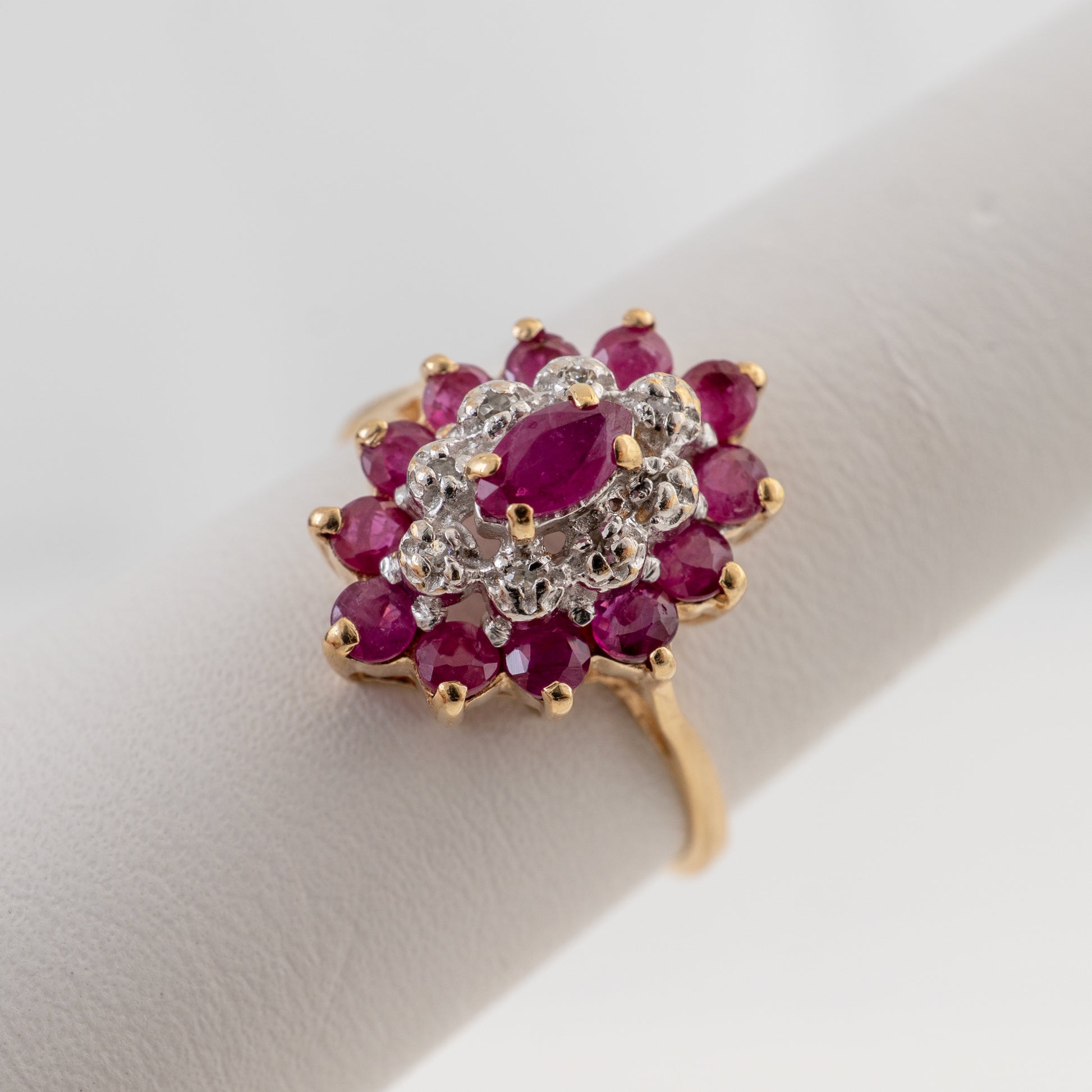 A yellow gold ring with a floral cluster setting featuring oval ruby gemstones and round clear stones, displayed on a white ring stand.
