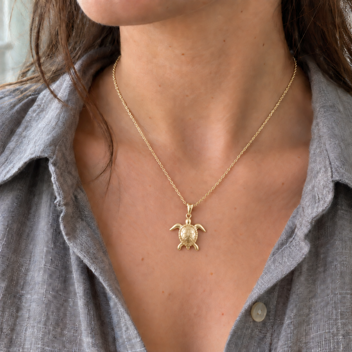 A close-up of a person wearing a silver sea turtle pendant necklace on a delicate chain, styled with a gray linen button-up shirt.