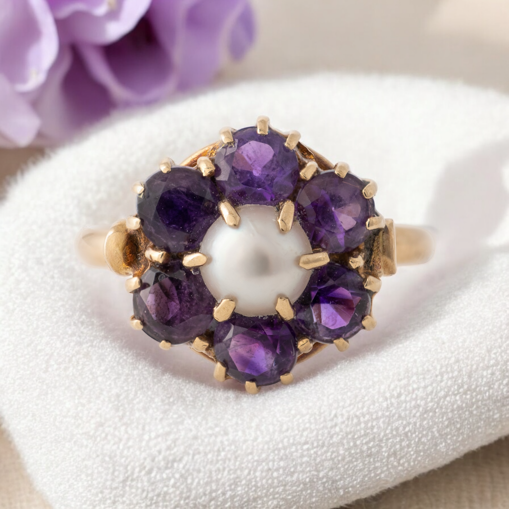 Vintage amethyst and pearl flower ring in 9ct yellow gold, displayed on a soft white cushion with a blurred purple flower background, showcasing its elegant 1950s design