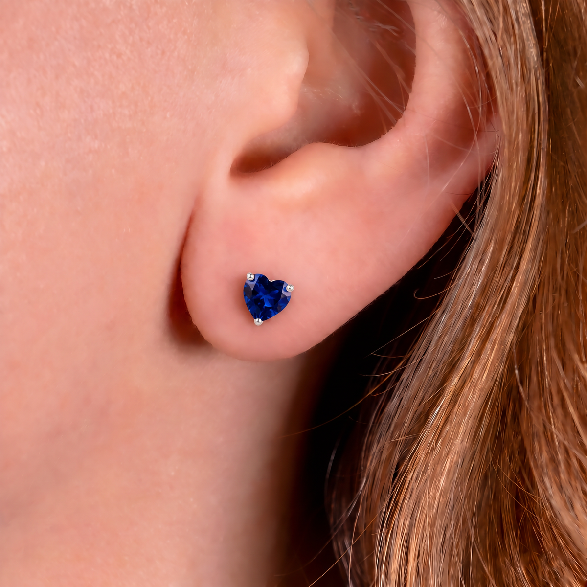 A close-up of a person's ear wearing a small heart-shaped ryal blue sapphire stud earring with tiny clear accent stones on a warm skin-toned background.