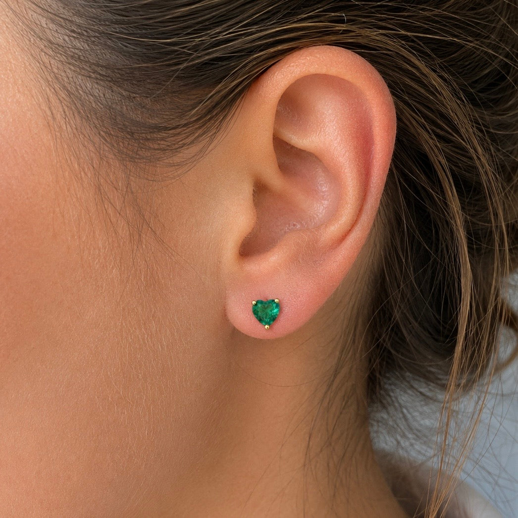 A close-up of a person's ear wearing a small heart-shaped green gemstone stud earring.