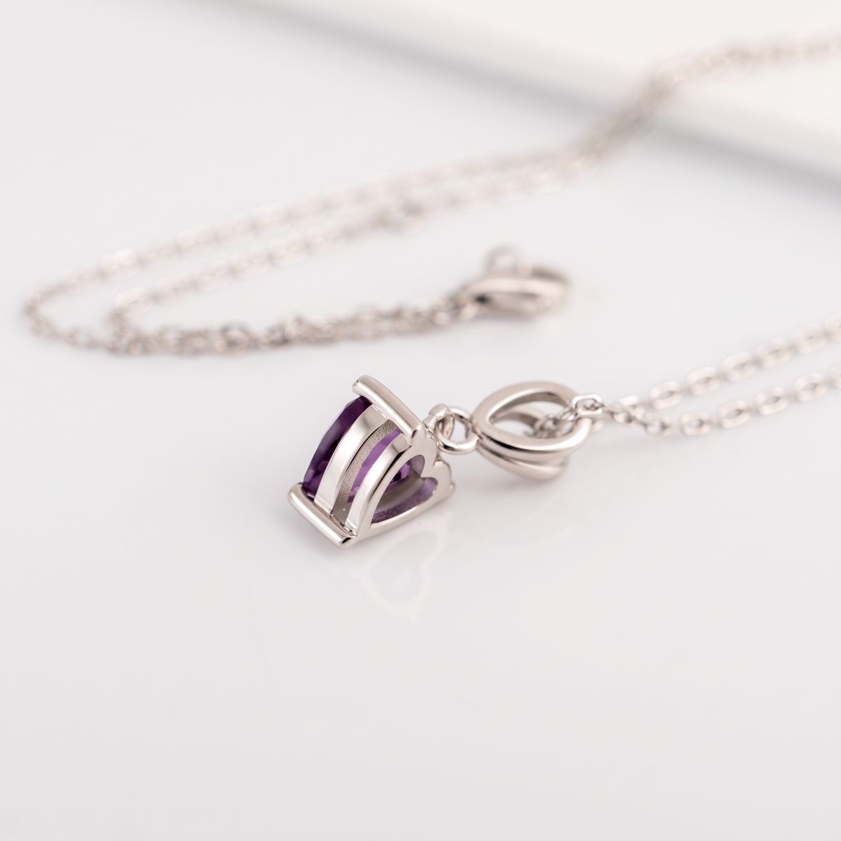 A silver pendant necklace featuring a small rectangular bezel-set purple amethyst heart, resting on a clean white surface with its matching chain blurred in the background.
