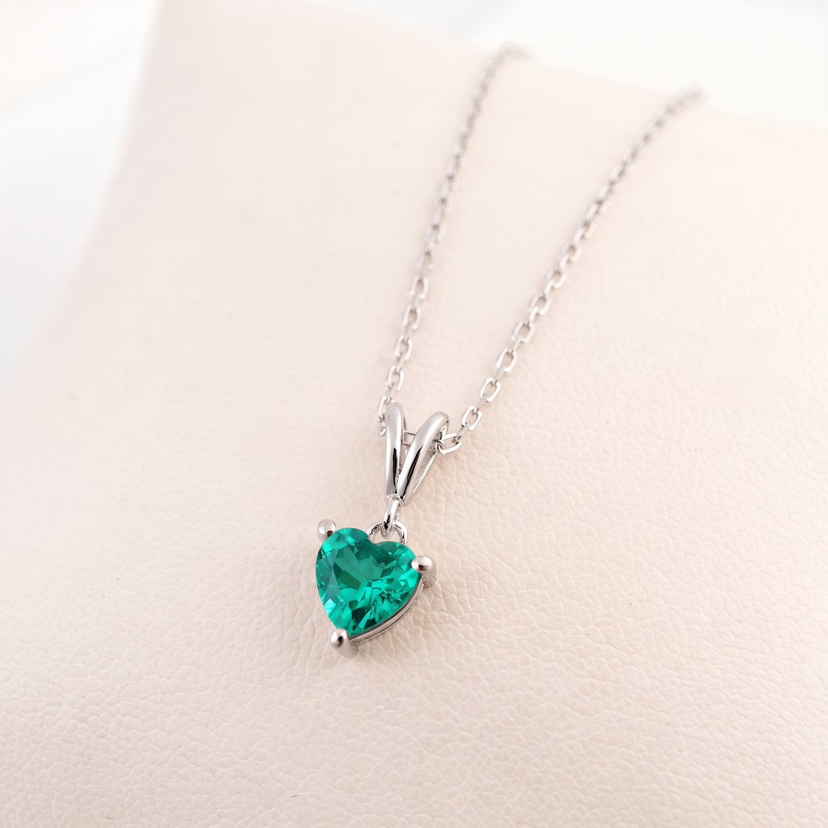 A heart-shaped emerald necklace pendant in a silver setting on a delicate silver chain, displayed on a light beige textured surface.