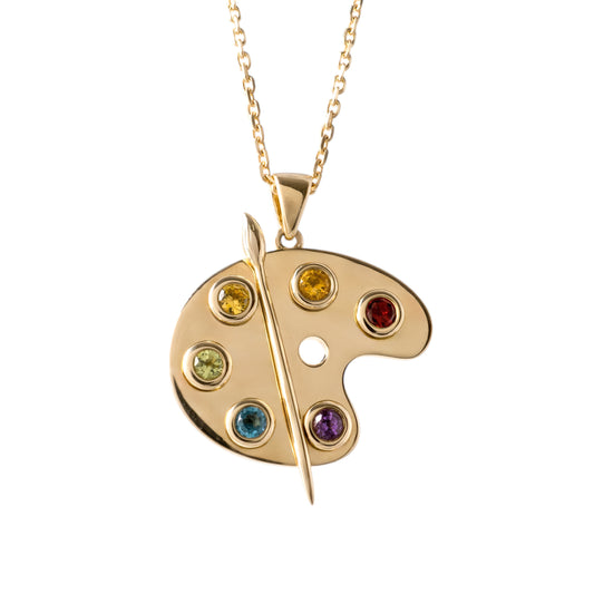 A gold artist palette pendant necklace featuring seven colorful gemstones in different hues, suspended from a delicate gold chain against a plain white background.