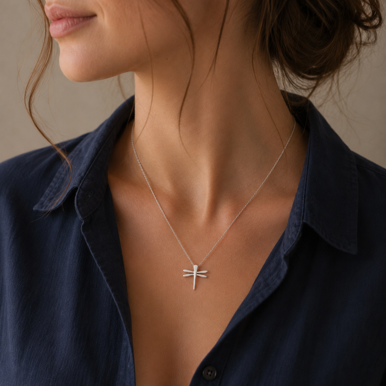 A close-up of a woman wearing a delicate silver dragonfly pendant necklace on a fine chain, paired with a dark navy blue collared shirt.