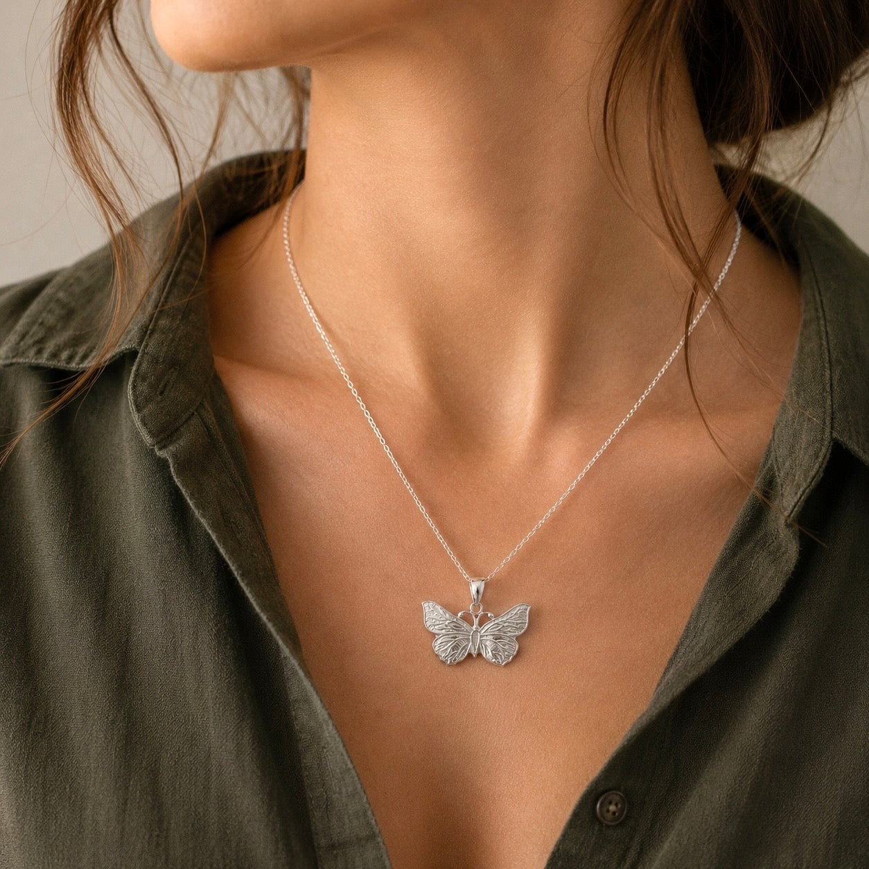 A close-up of a model wearing a delicate silver butterfly pendant necklace on a fine chain, styled with an olive green shirt.