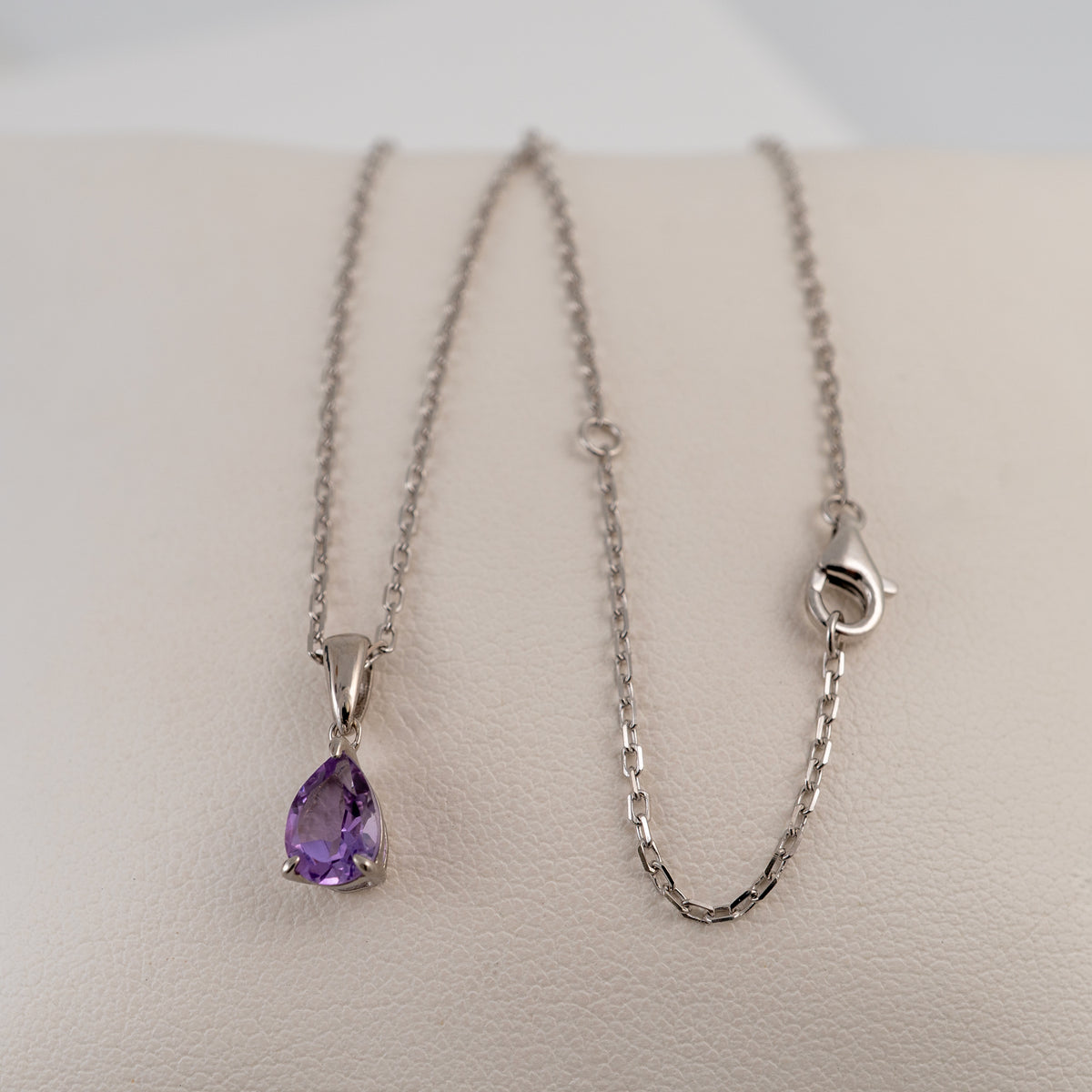 Sterling silver chain necklace with a teardrop-shaped amethyst pendant displayed on a light beige textured surface