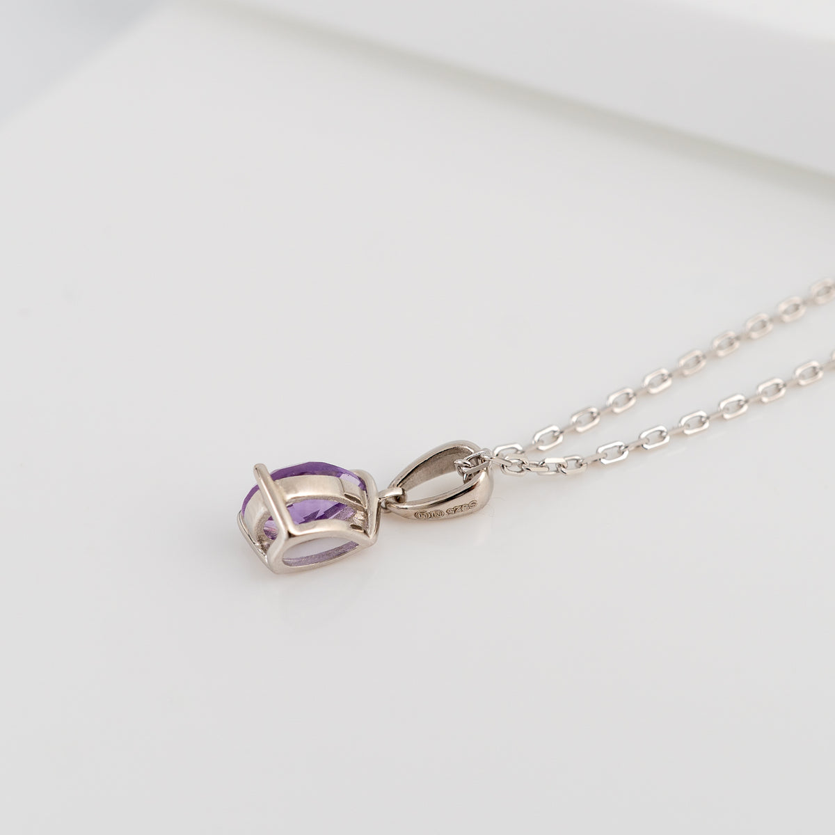 A silver pendant necklace featuring a small oval purple gemstone in a geometric prong setting, resting on a clean white surface.