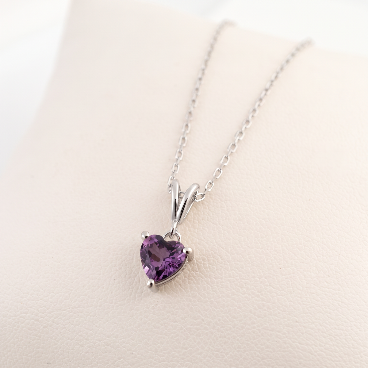 A silver heart-shaped pendant necklace with a purple gemstone, displayed on a light beige textured surface.