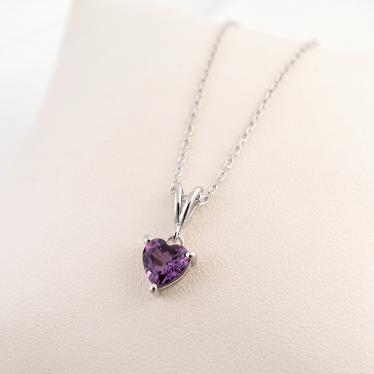 A silver heart-shaped pendant necklace with a purple gemstone, displayed on a light beige textured surface.