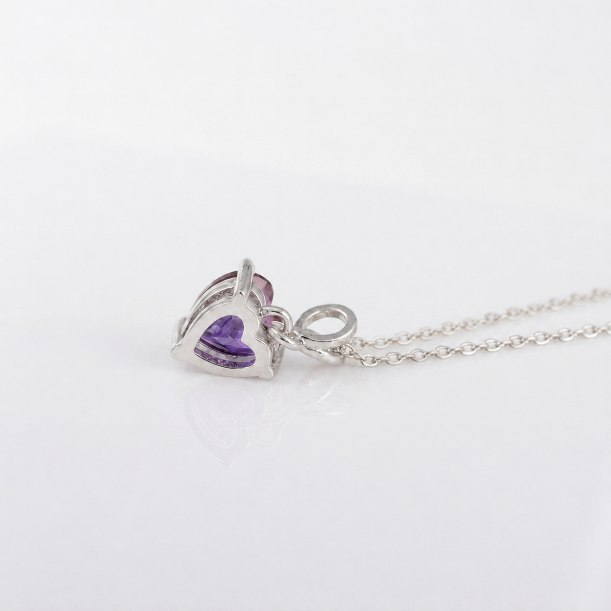 A silver heart-shaped pendant necklace featuring a purple amethyst gemstone, displayed on a clean white reflective surface.