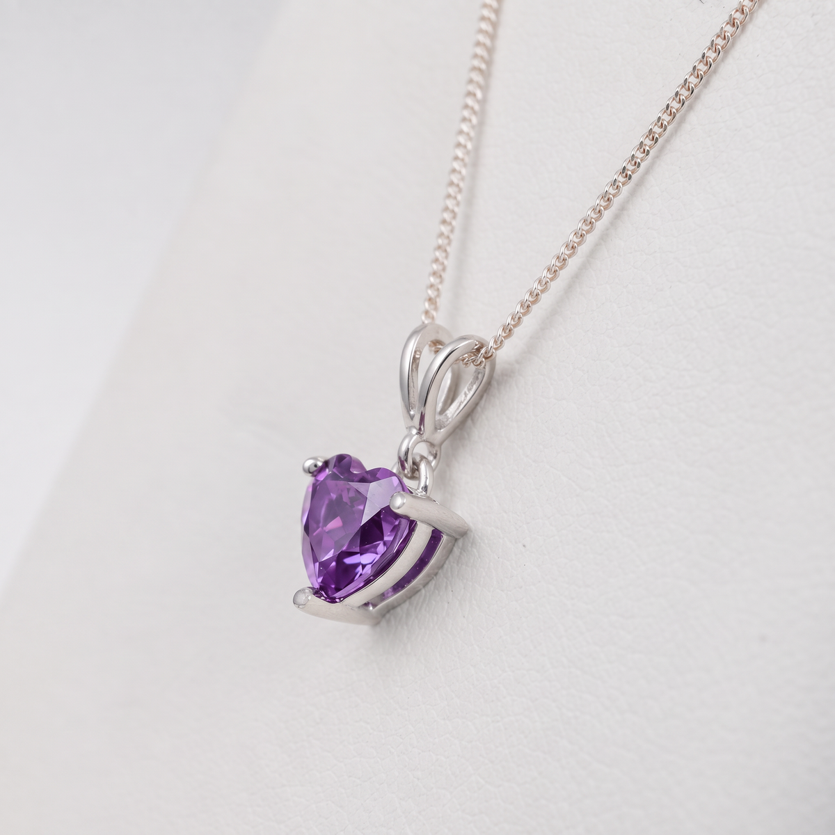 A heart-shaped amethyst pendant in a silver-colored setting on a delicate silver chain, displayed on a white background.