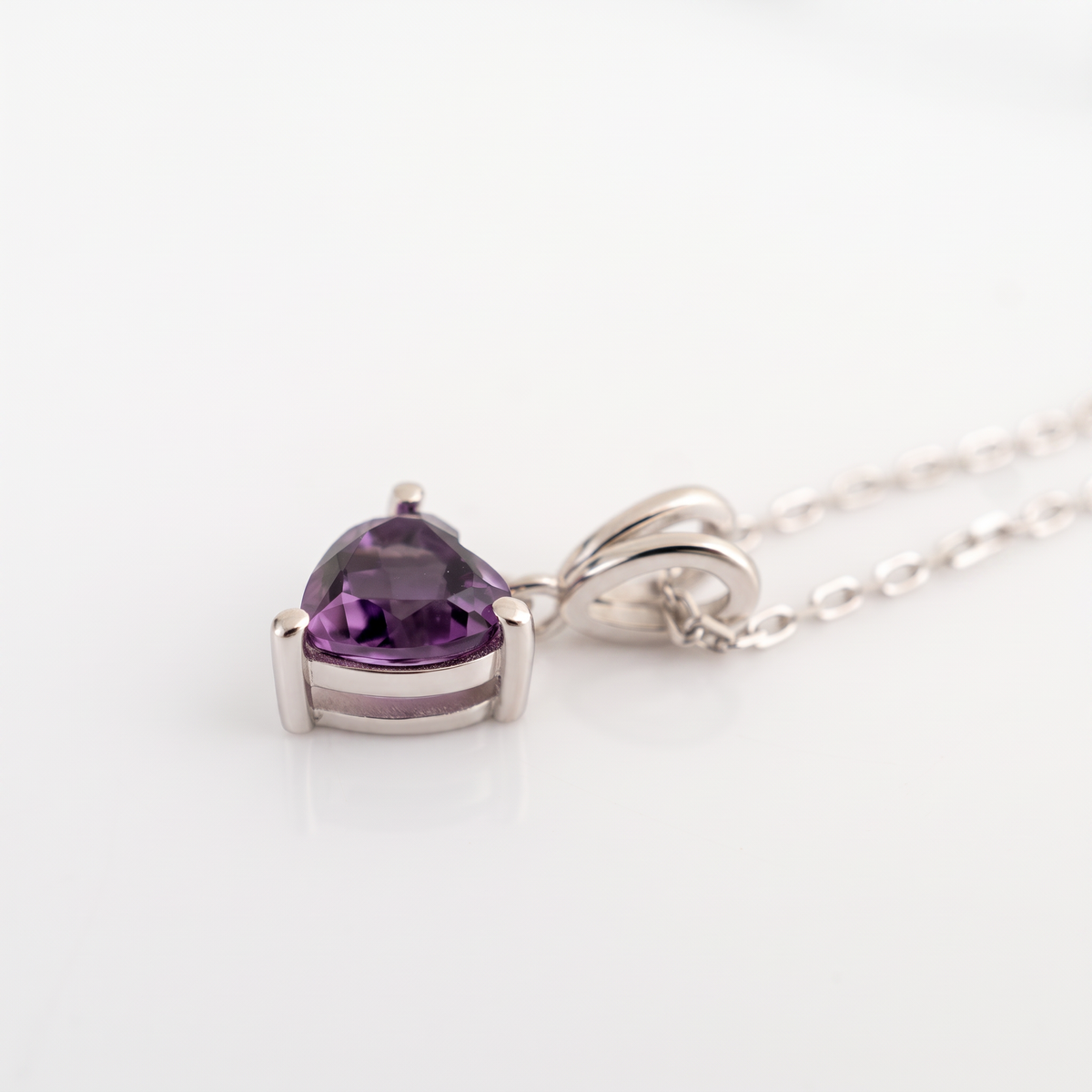 A heart-shaped purple amethyst gemstone pendant in a silver setting on a delicate silver chain, photographed on a clean white background.