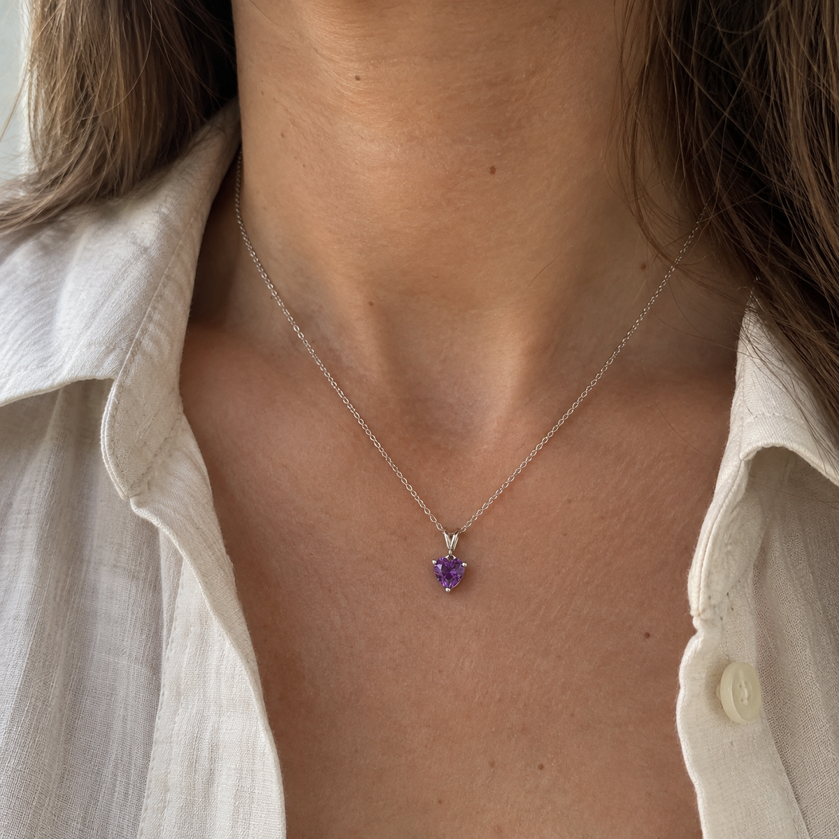 A close-up of a person wearing a delicate silver chain necklace with a small heart-shaped purple gemstone pendant, styled with a light-colored linen shirt.