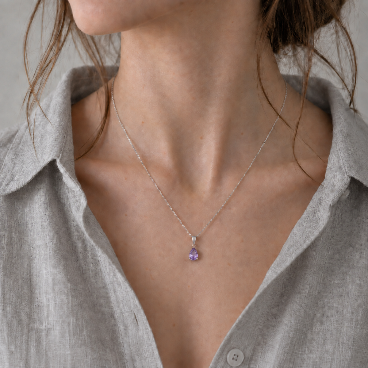 A close-up of a model wearing a delicate silver chain necklace with a small lavender-amethyst teardrop pendant, styled with a light gray linen shirt.