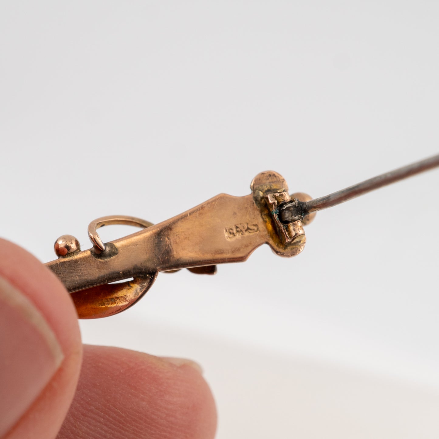Close-up of a gold-toned pin or clasp being held between fingers, showing engraved markings and a thin metal post extending to the right against a plain light background.