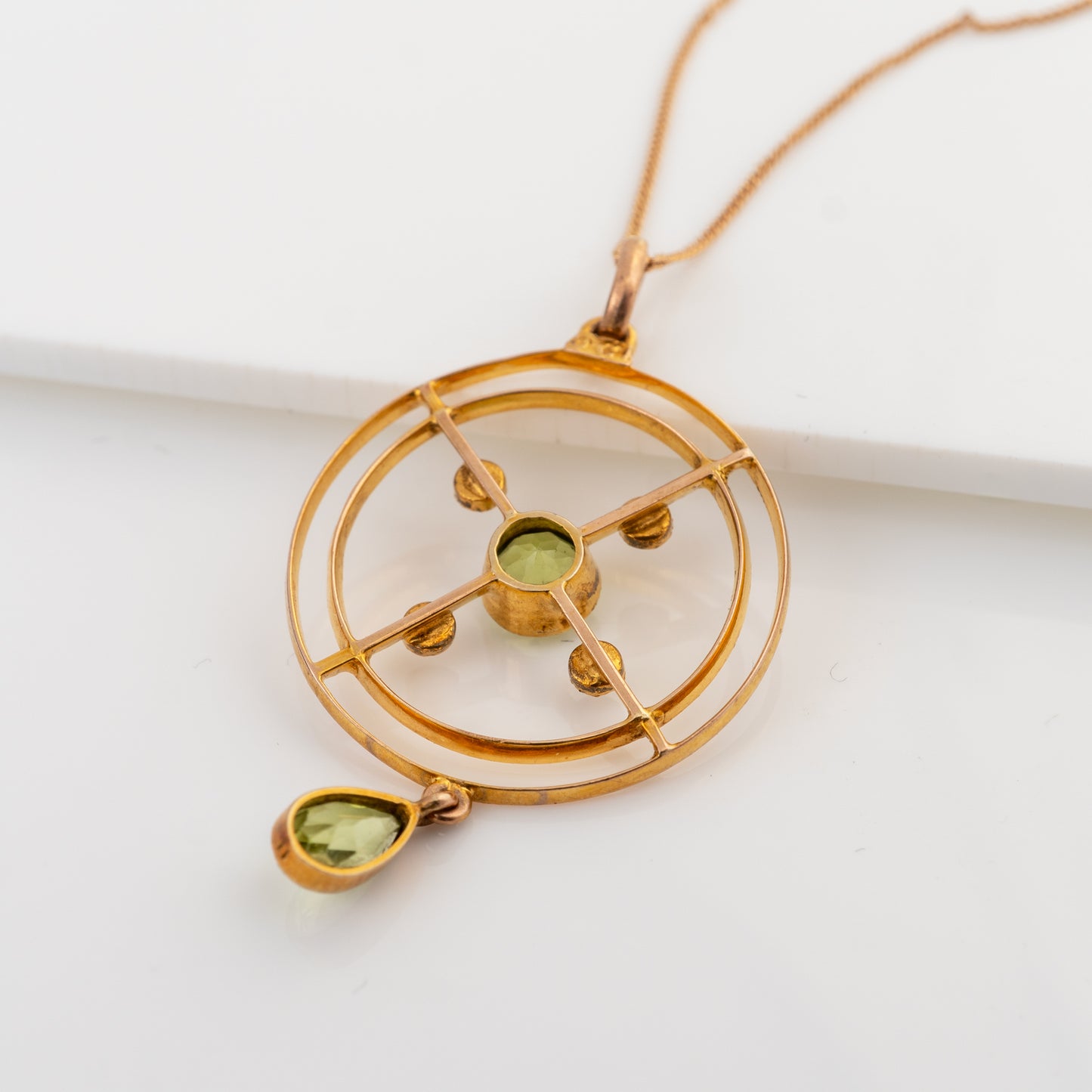 A gold-toned geometric pendant necklace featuring concentric circular rings, a central green gemstone, and a dangling teardrop-shaped green stone, displayed on a clean white surface.